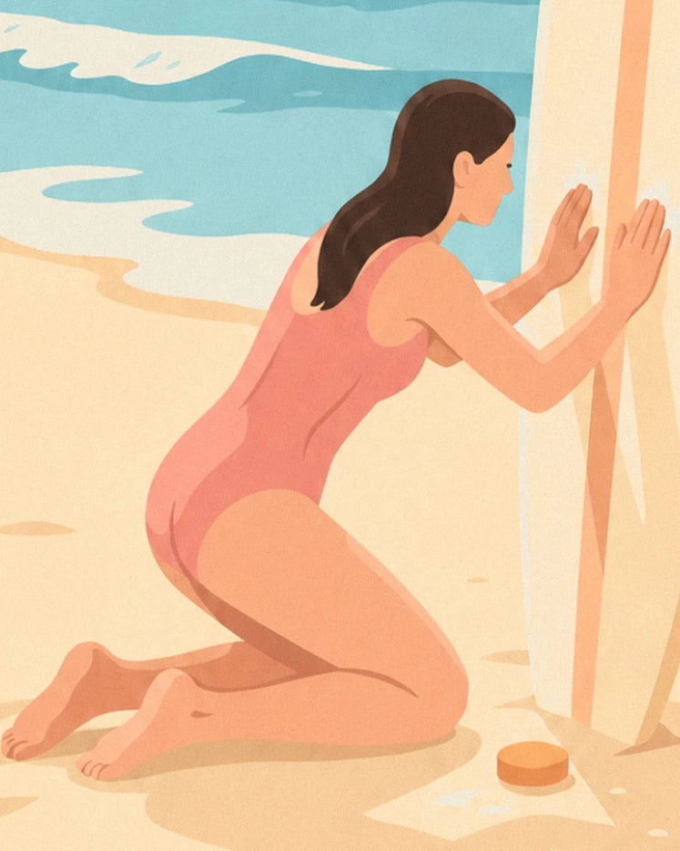 The surfer in a coral swimsuit kneeling in front of her board, with her bar of wax on the sand